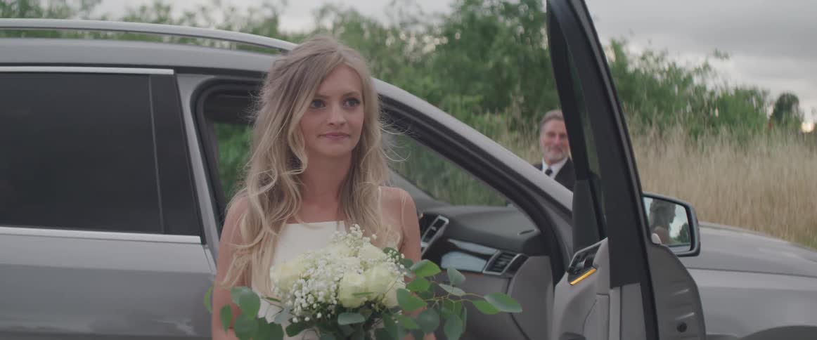 A woman stepping out of a vehicle with a white dress and a bouquet with a man in a suit in the background