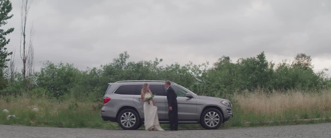 A woman with a white dress and a bouquet and a man with a suit cross the street with a gray truck in the background