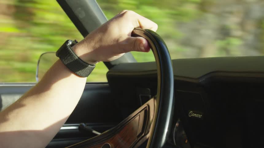 A man wearing a watch on his left wrist drives a classic car on a hillside road