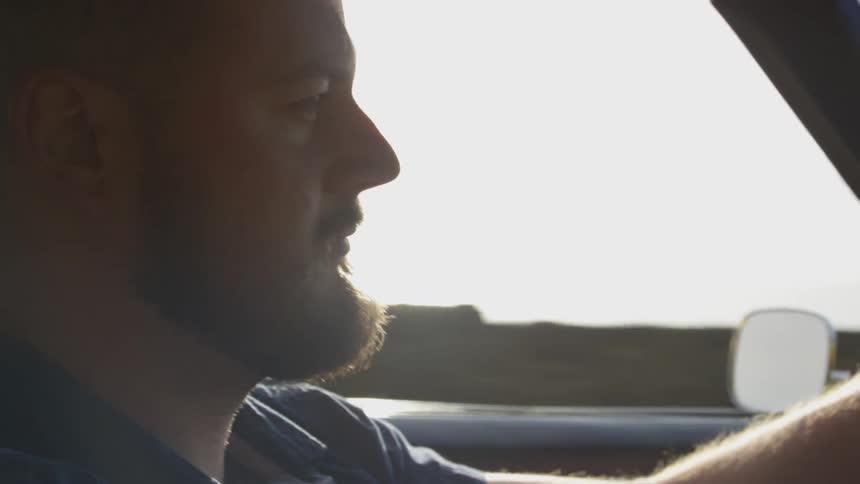 A bearded man driving a vehicle down a road on the coast