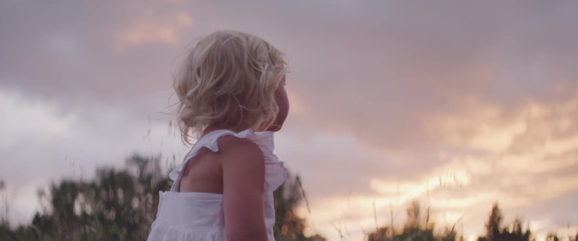 Small blonde haired girl in white summer dress turns around towards the trees at the edge of a field