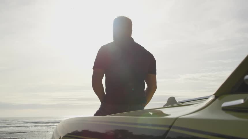 A man sits on the bonnet of his car which has a racing stripe down the side and is parked on a coast location