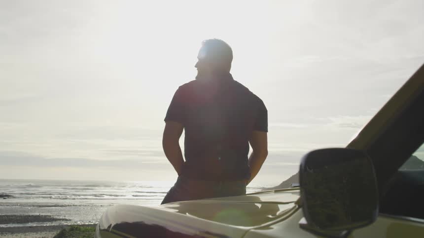 Man leaning on the hood of his car on the beach and looks out over the ocean at the horizon