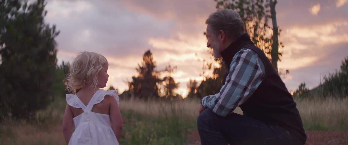 An older white man and a young white girl look at the sunset in a field