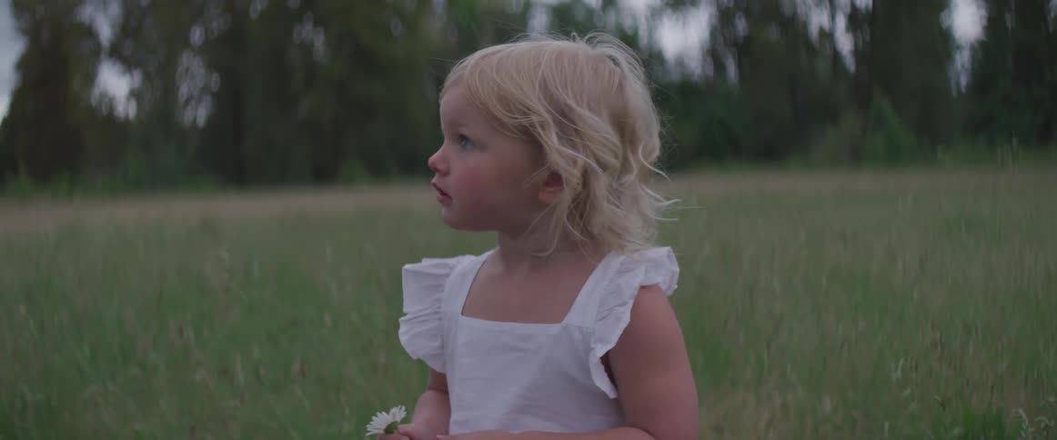 A girl with blonde hair holds a flower as she walks in the grass