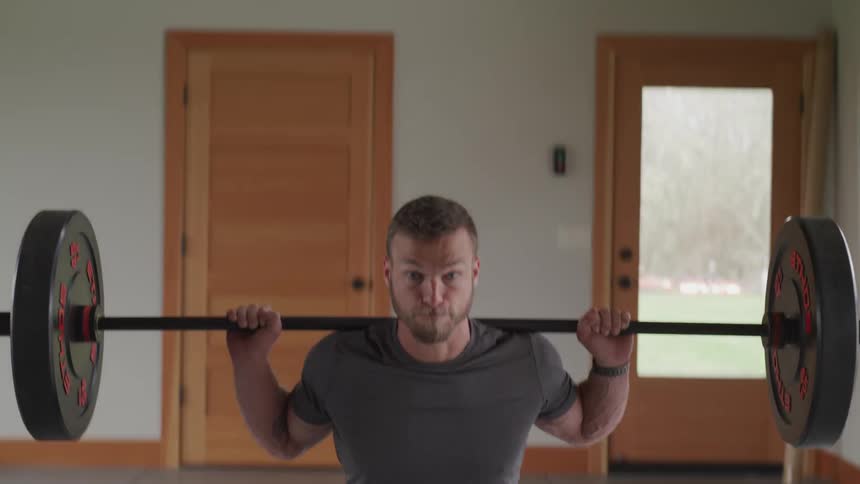 Bearded blonde man does a barbell squat indoors during daylight