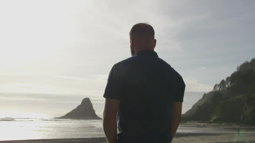 A man in a polo stands with his arms at his sides and gazes at the ocean