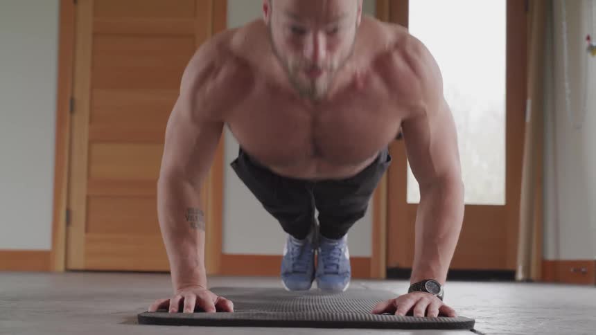 Shirtless man performs series of pushups on a mat while working out in a room in the afternoon