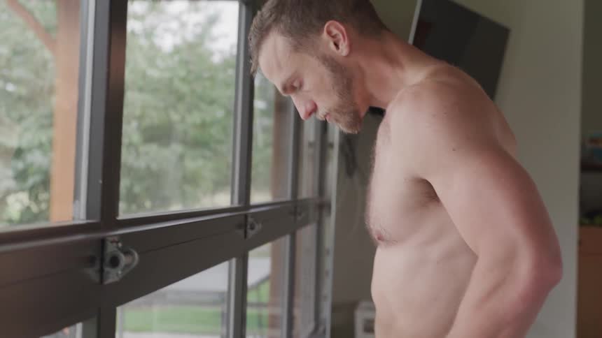 A shirtless young white man catching his breath near a window