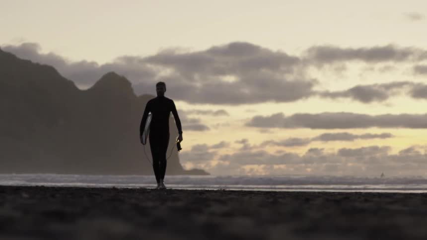 A surfer carries his board as he walks along the shoreline at sunset.
