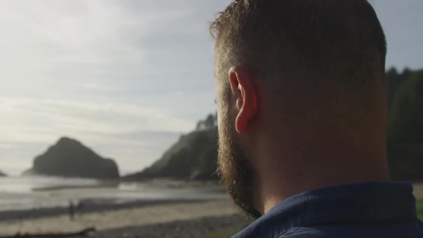 A bearded man stands on a beach and looks out at the waves and large rocks protruding above the ocean surface