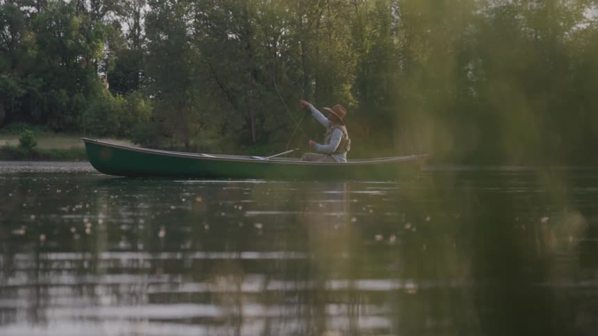A man sitting in a green boat throws a fishing lure into the water