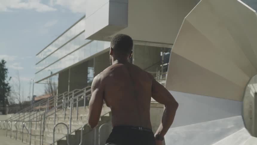 A shirtless man runs outdoors past a building and steps
