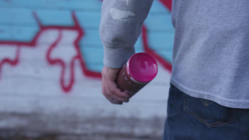 Person in a sweater walks up to a wall with a bottle of spray paint
