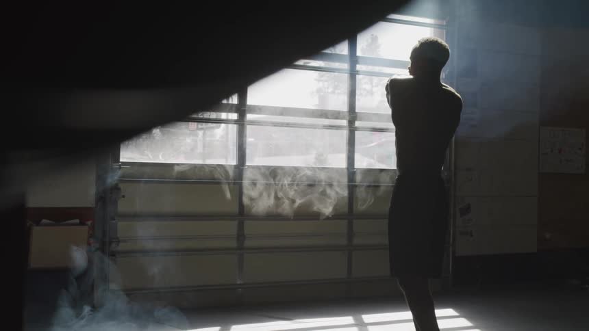 A shirtless male twists his body and stretches out his arms in a gym in front of some windows