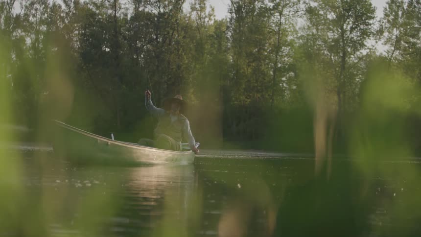 A fisherman sits in a boat and casts his fishing pole