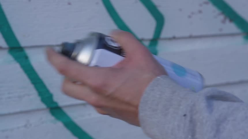 White person is shaking a spray paint can in front of a white siding wall with green marks on it
