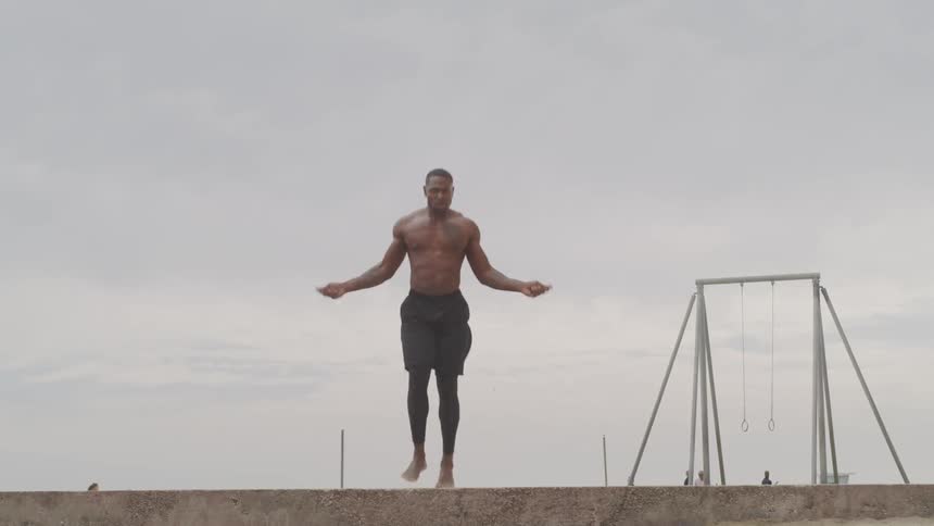 Shirtless muscular black man jumps rope on the beach on a cloudy day