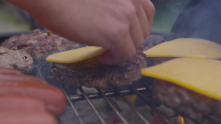 A persons hand as they place cheese on burgers cooking on a smoking grill