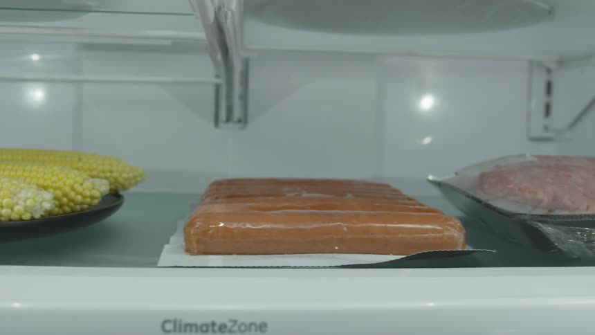 Food sitting on a shelf in a refrigerator includes corn on the cob hot dogs and ground beef