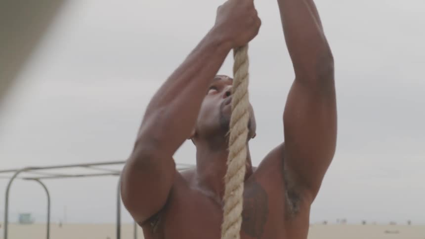 A black man climbing a rope for exercise at the beach