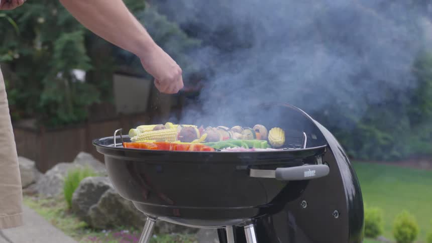 A man sprinkles salt on vegetables cooking on an outdoor charcoal grill