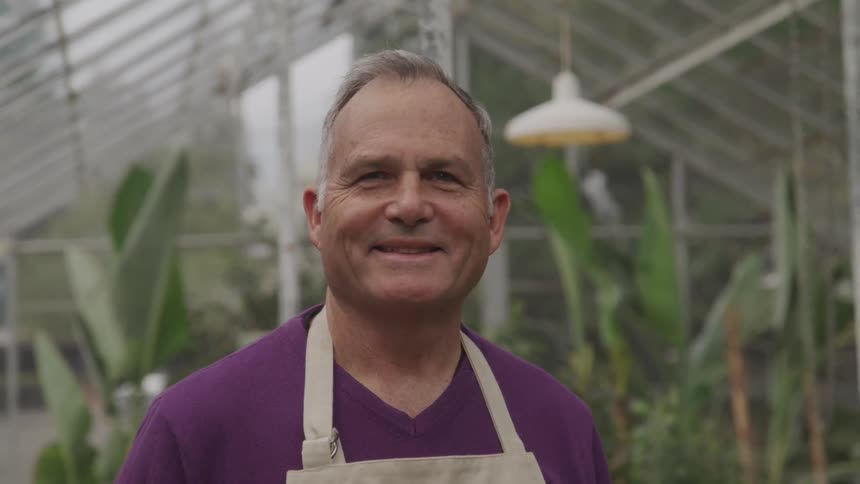 A man wearing an apron smiles at the camera while standing in a greenhouse