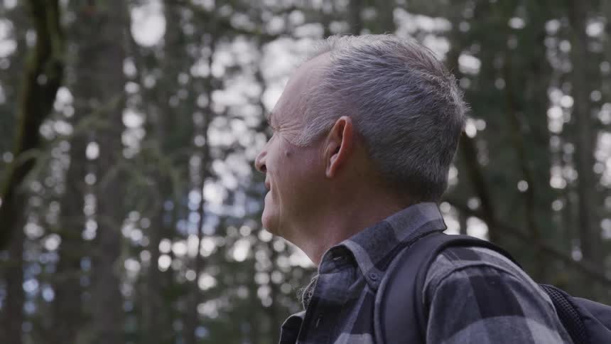 Old man wearing backpack looks ahead and smiles in the woods during daylight
