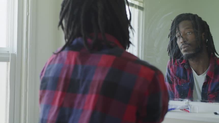 A man with dreads looks up and into the bathroom mirror