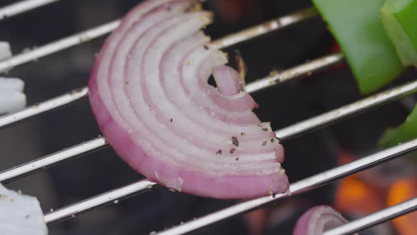 A purple onion sits upon a metal rack above a grill where it is surrounded by other other onions and bell peppers