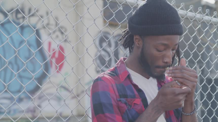 A black man lighting a cigarette while leaning against a chain link fence