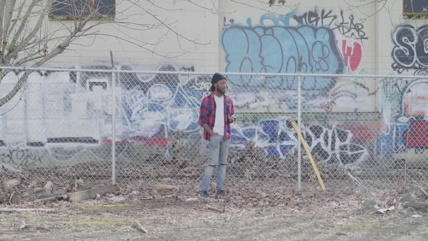 A black man stands and smokes in front of a fence thats near a graffiti covered wall