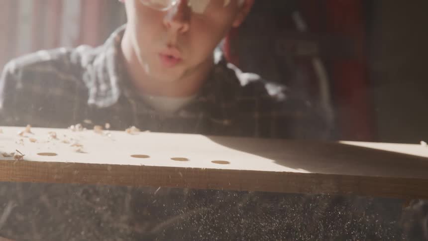 A man lifting a wooden board from a drill press and blowing the sawdust off the top