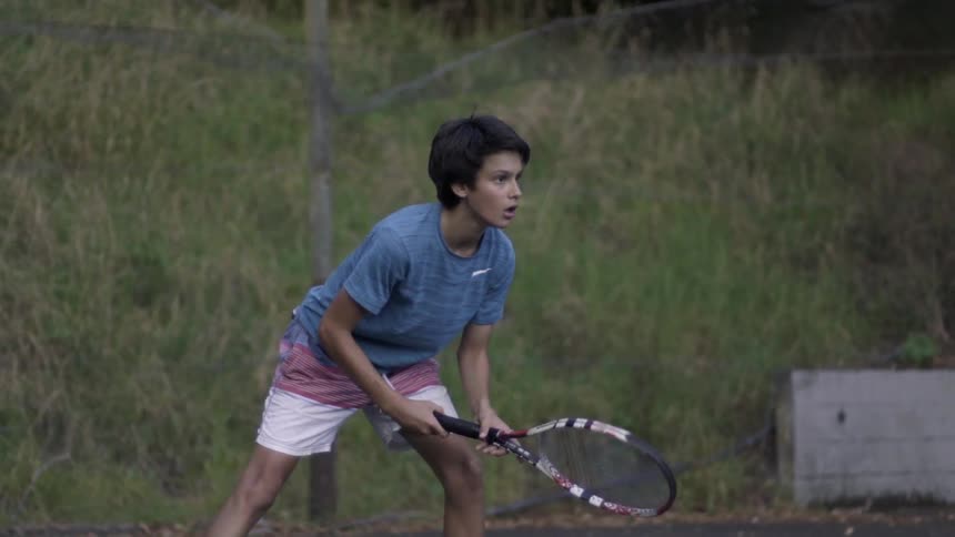 A boy is playing tennis on a court with a hill behind him