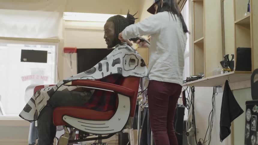 A black man getting his hair cut by a white woman in a ball cap in a barber shop during the day