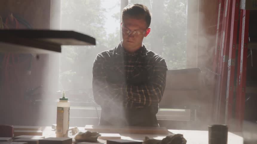 A man in a plaid shirt and safety glasses stands at a desk in front of a window