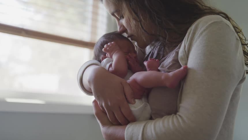 A woman cradles and kisses a newborn while standing next to a window