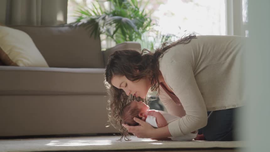 A woman with long brown hair leans down to kiss a small infant on the floor of a living room