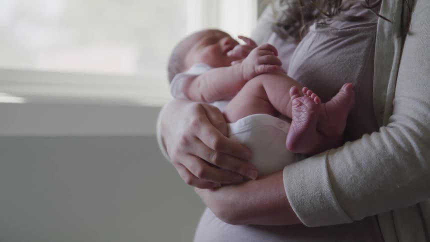A woman holds a very small baby near a window as the baby is wiggling and squirming