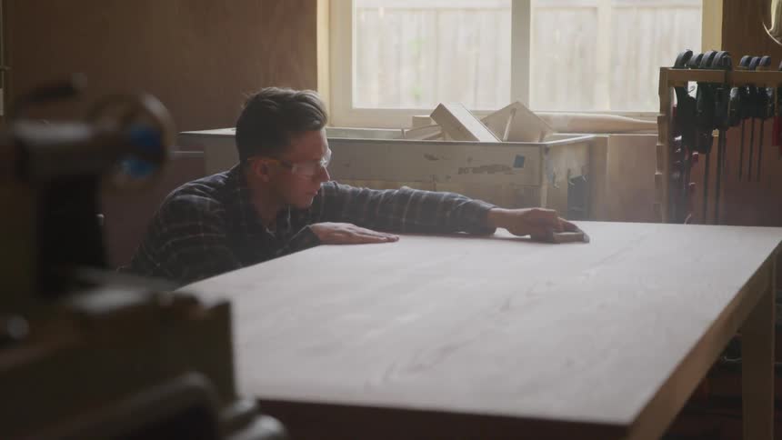 A man wearing goggles sands a wood table
