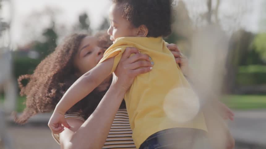 A female is lifting a young boy off a swing at a playground and then holds him in her arms as he points at something