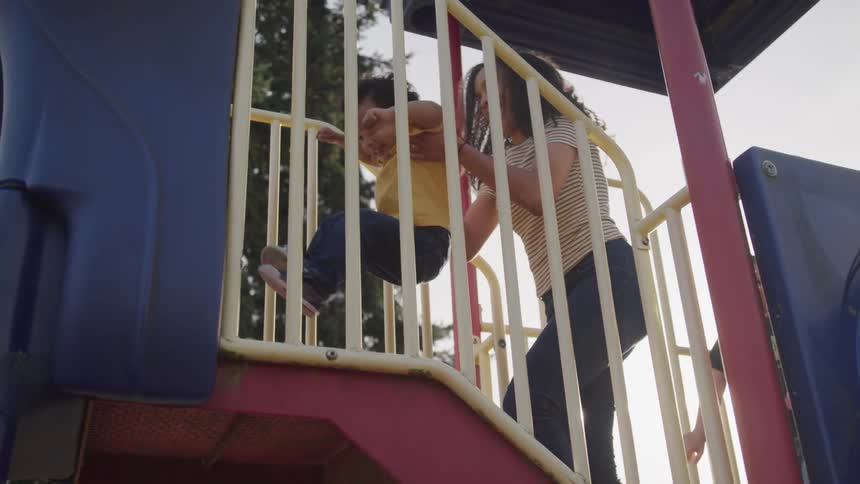 A mother lifts her son up to ride a slide at the playground