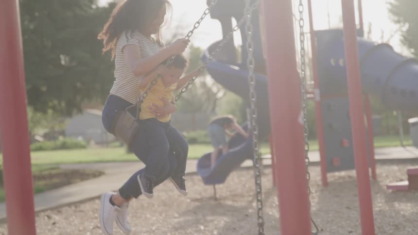 A mother and son swing on a park swing set on a sunny summer day