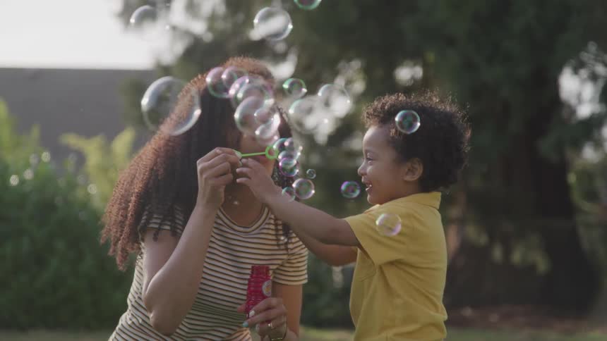 Mother is blowing bubbles and she laughs as the child tries to grab the wand from her