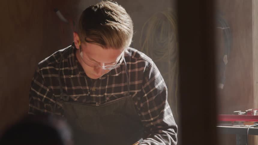 A man in a plaid shirt safety glasses and apron focuses intently on what he is writing