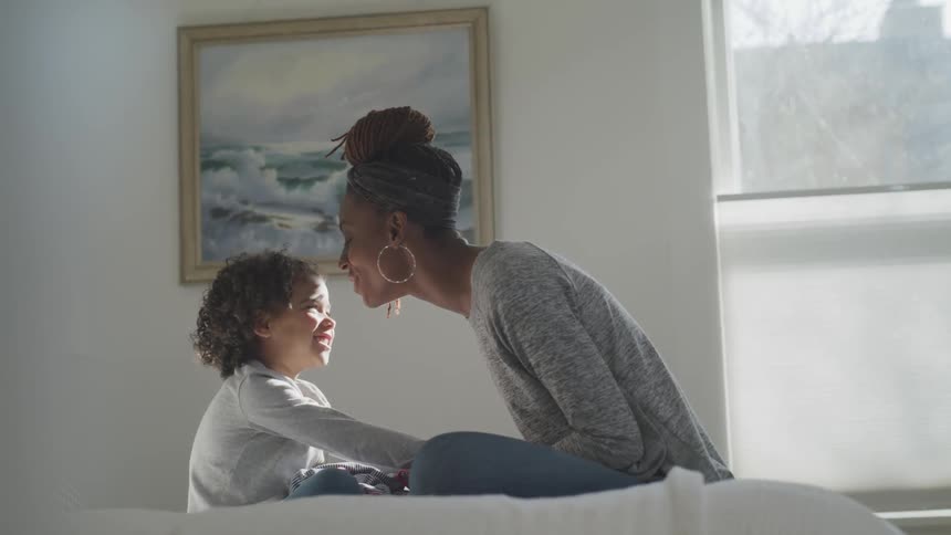A woman and child sit on a bed together and chat and laugh and give kisses