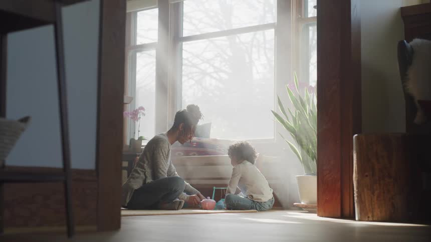 Mother and child sitting on the floor next to a large window having a tea party
