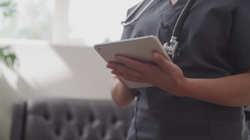 A medical professional wearing gray scrubs has a stethoscope around her neck and is entering information into a tablet