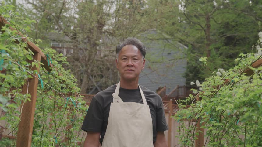A man wearing an apron stands outdoors near plants