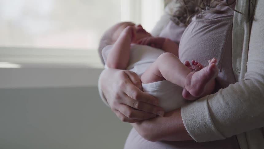A woman standing near a window holding a crying baby